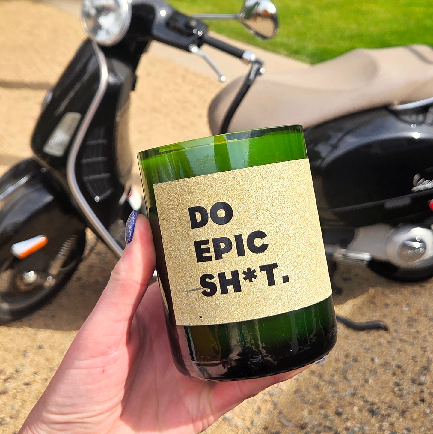 Person holding a green wine bottle candle with a sparkly gold label saying "do epic sh*t", in front of a vespa.