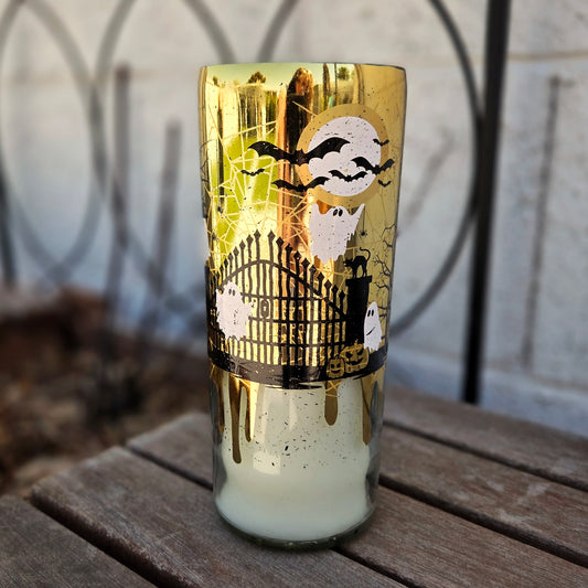 Front-facing view of a gold wine bottle candle wrapped in eerie Halloween art with ghosts, a haunted gate, bats, and a glowing moon. A sustainable, hand-poured candle with coconut soy wax and a wooden wick.