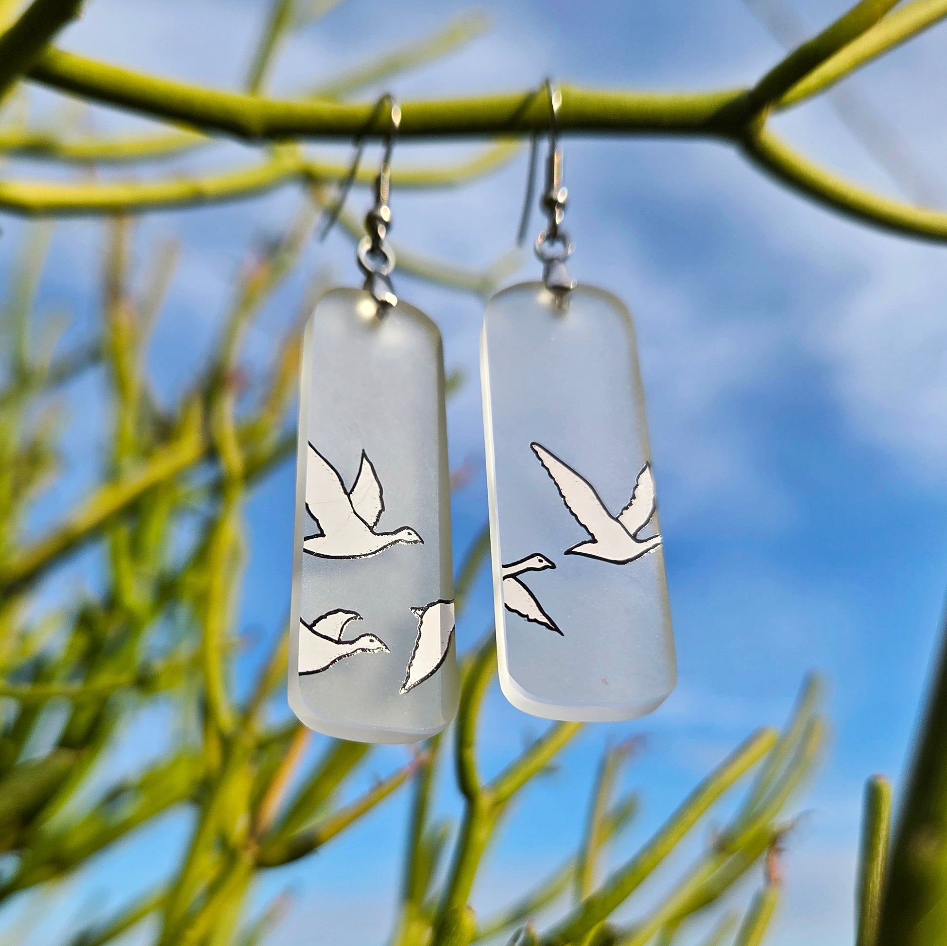 A pair of rectangular frosted glass drop earrings with gray goose silhouettes, hanging from silver fishhook wires against a blurred background of green tree branches and blue sky.