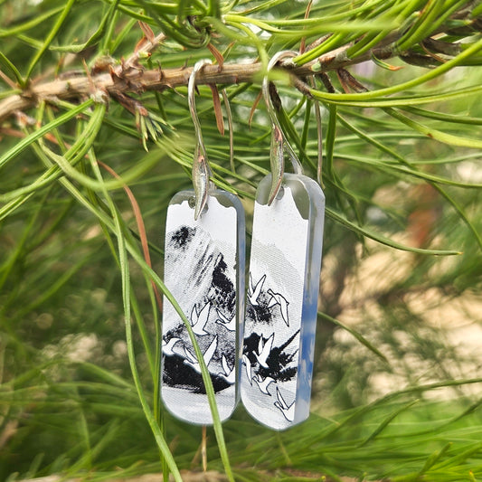Pair of glass earrings with black and white nature designs hanging from a branch with green leaves.