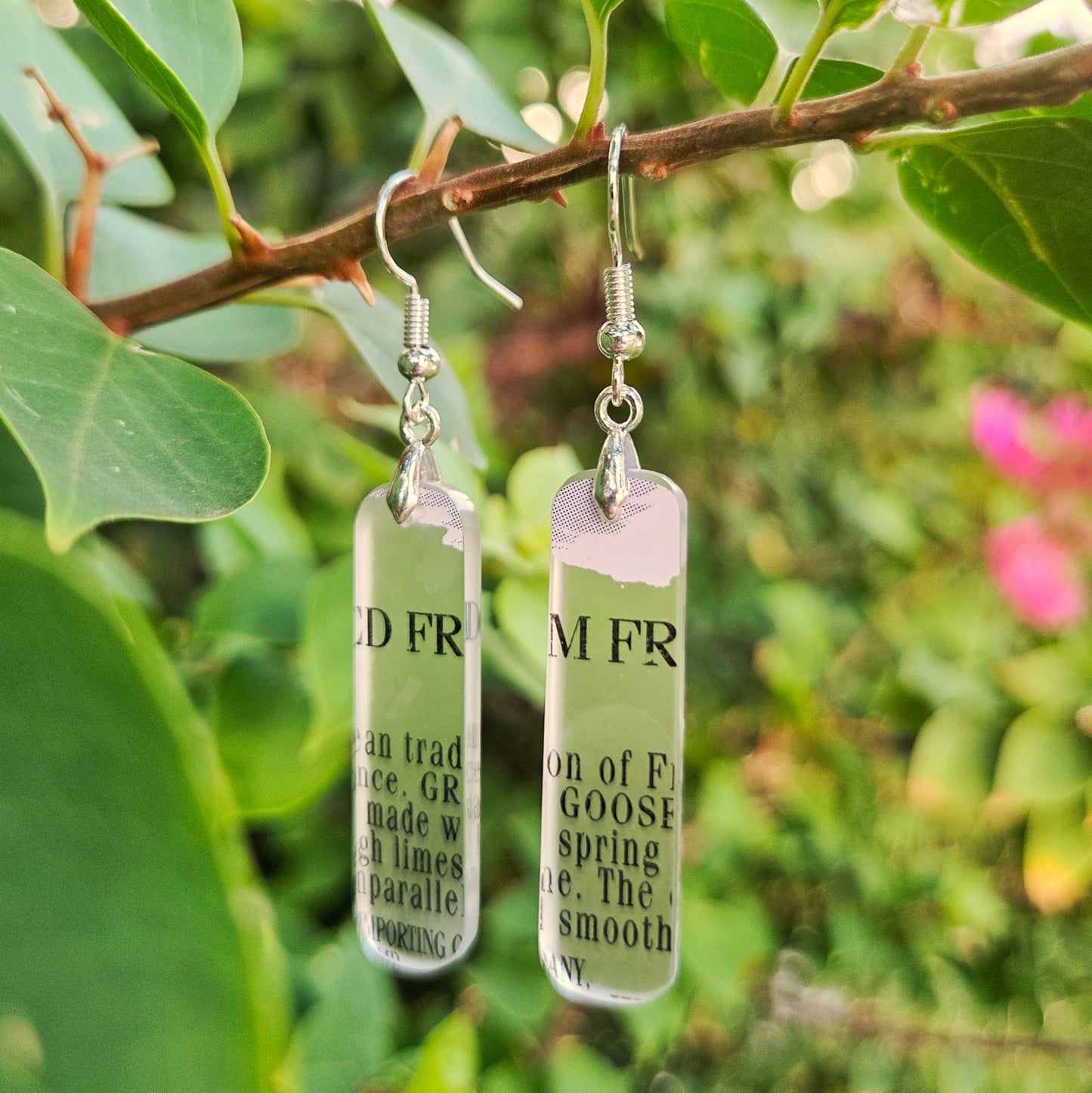Two rectangular clear glass drop earrings with silver fishhook wires and printed black text, hanging from a tree branch with green leaves and blurred pink flowers in the background.