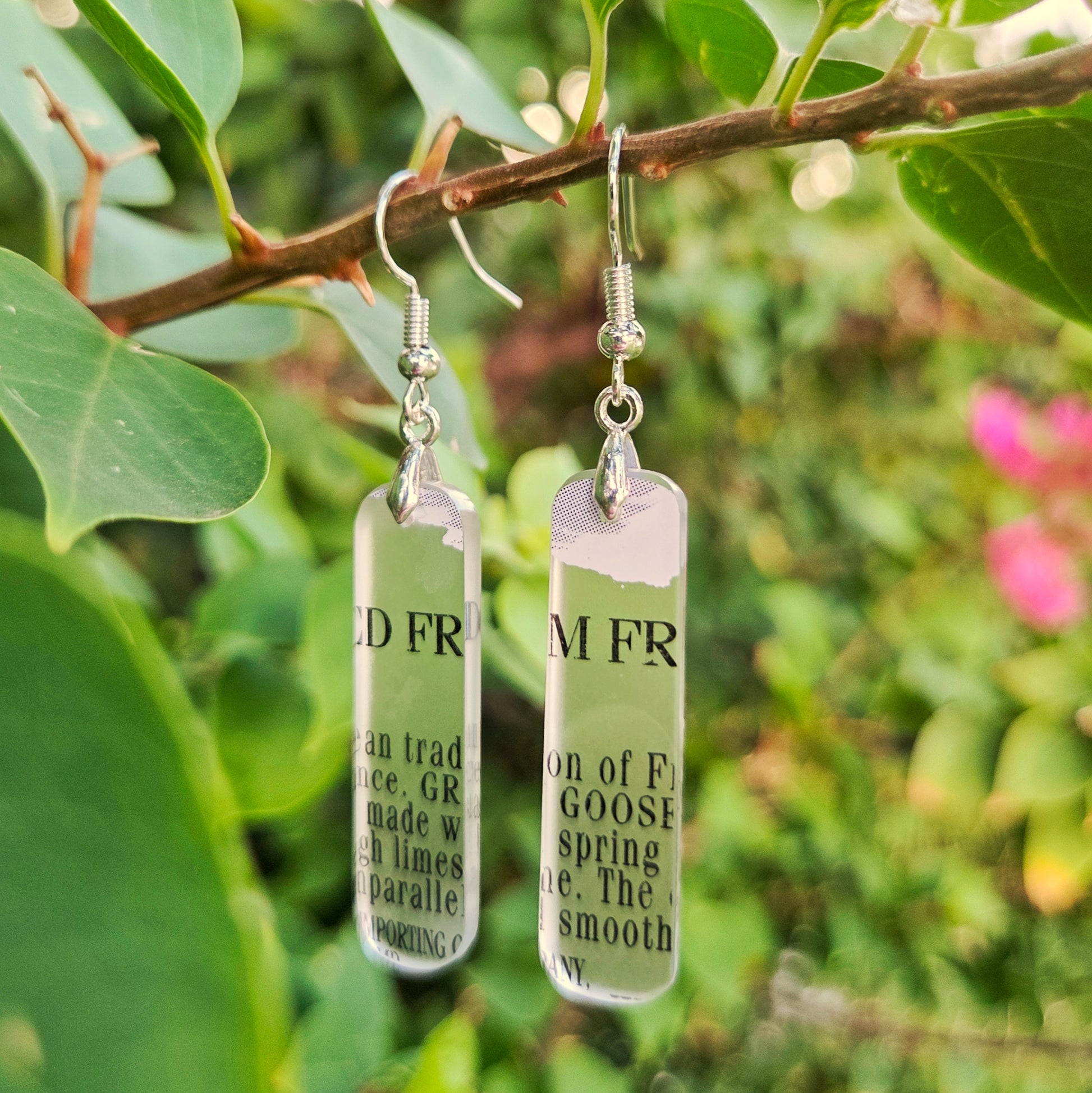Two rectangular clear glass drop earrings with silver fishhook wires and printed black text, hanging from a tree branch with green leaves and blurred pink flowers in the background.