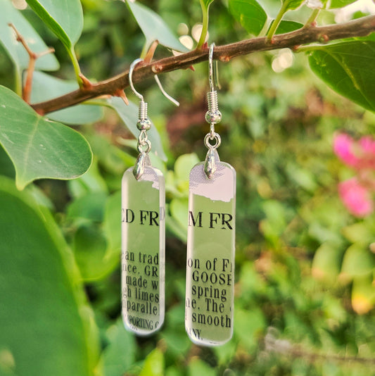 Two rectangular clear glass drop earrings with silver fishhook wires and printed black text, hanging from a tree branch with green leaves and blurred pink flowers in the background.