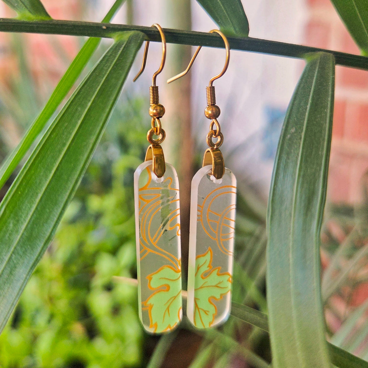 A pair of rectangular dangling earrings with a frosted light green and white background, decorated with gold leaf patterns, hanging from gold fishhook wires. The earrings are photographed hanging from a green palm branch with a blurred garden background.