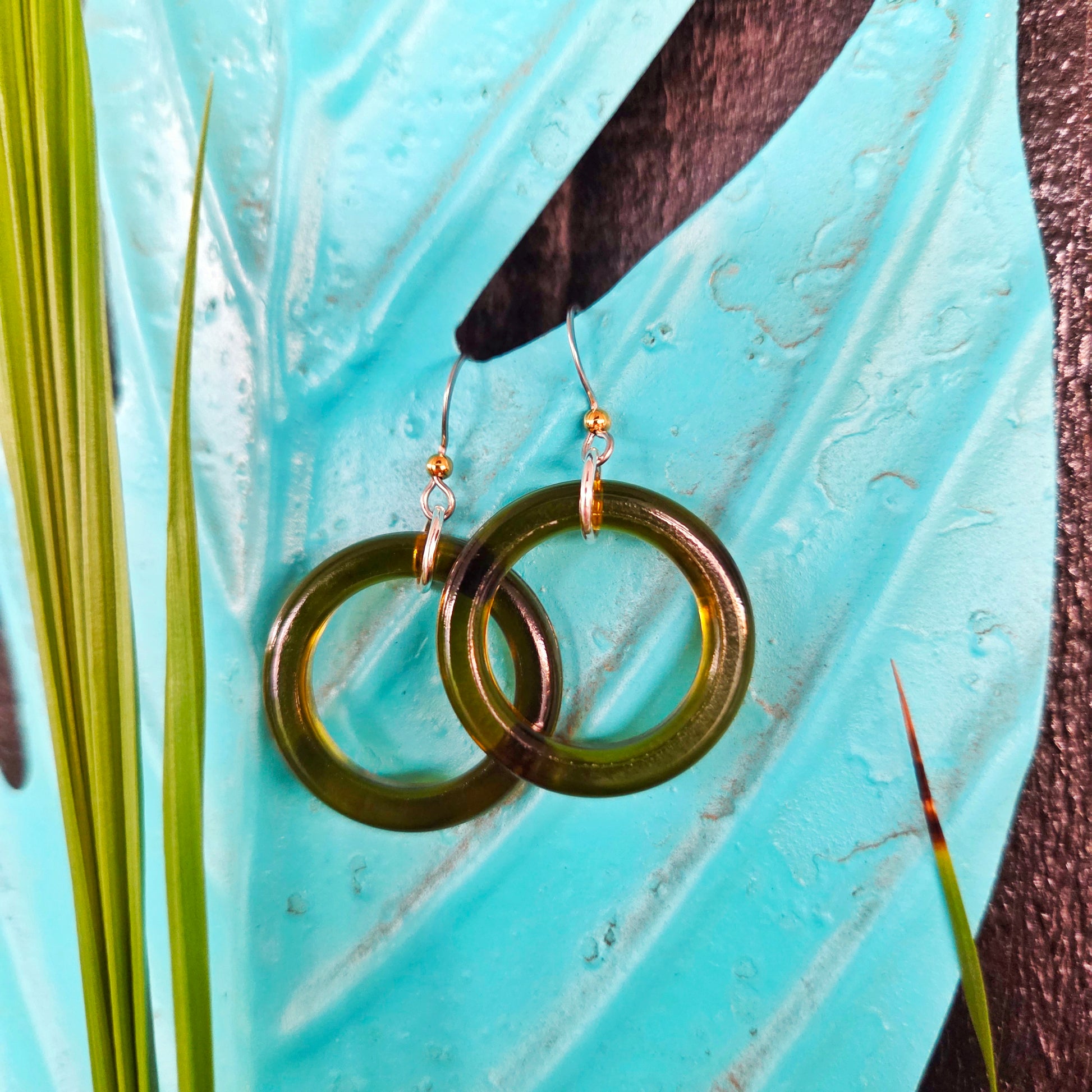 Two olive green translucent glass hoop earrings with silver-toned hooks, displayed on a glossy turquoise leaf with green plant stems.