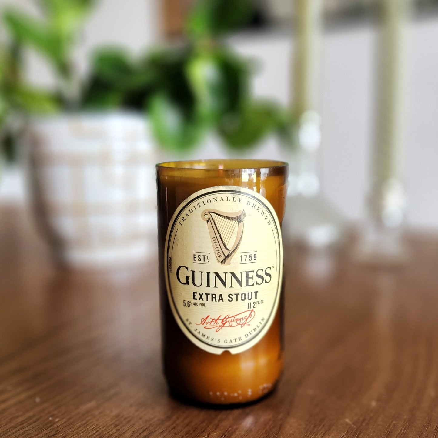 A hand-poured coconut soy wax candle crafted from a recycled Guinness Extra Stout beer bottle. The classic beige and black label features the iconic Guinness harp, adding a timeless Irish touch. This eco-friendly wooden wick candle has a polished glass rim, highlighting expert craftsmanship. Placed on a wooden surface with a softly blurred background of greenery and elegant candlesticks, this upcycled beer bottle candle is perfect for Guinness fans, craft beer lovers, and sustainable shoppers.