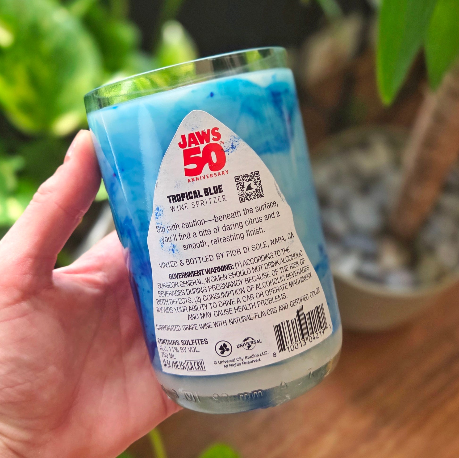 Back view of a Jaws-themed wine bottle candle featuring the original Tropical Blue wine spritzer label. The candle is set in a recycled glass bottle with swirled ocean-blue wax, highlighting its handmade, upcycled appeal.
