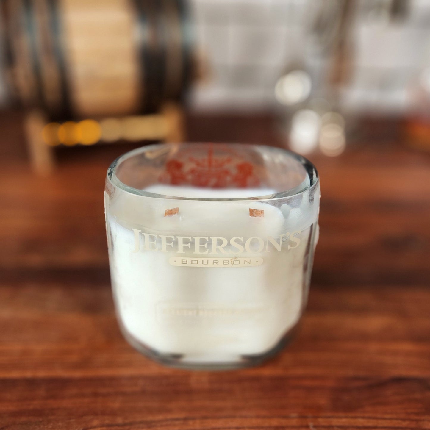 A hand-poured coconut-soy candle in a recycled Jefferson's Bourbon glass. Features double wooden wicks and the original etched design. Displayed on a wooden surface with a blurred barrel in the background, combining elegance and eco-friendliness.