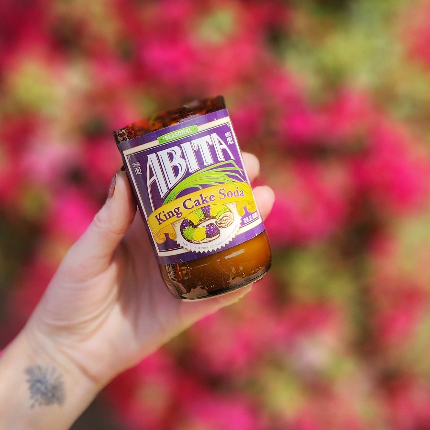 A hand-poured coconut soy wax candle made from a recycled Abita King Cake Soda bottle. The label showcases vibrant Mardi Gras colors with an illustration of a classic King Cake, celebrating New Orleans' festive spirit. This eco-friendly wooden wick candle has a polished glass rim, highlighting expert craftsmanship. Held against a blurred backdrop of bright pink bougainvillea, this upcycled soda bottle candle is a unique and sustainable gift for Mardi Gras enthusiasts.