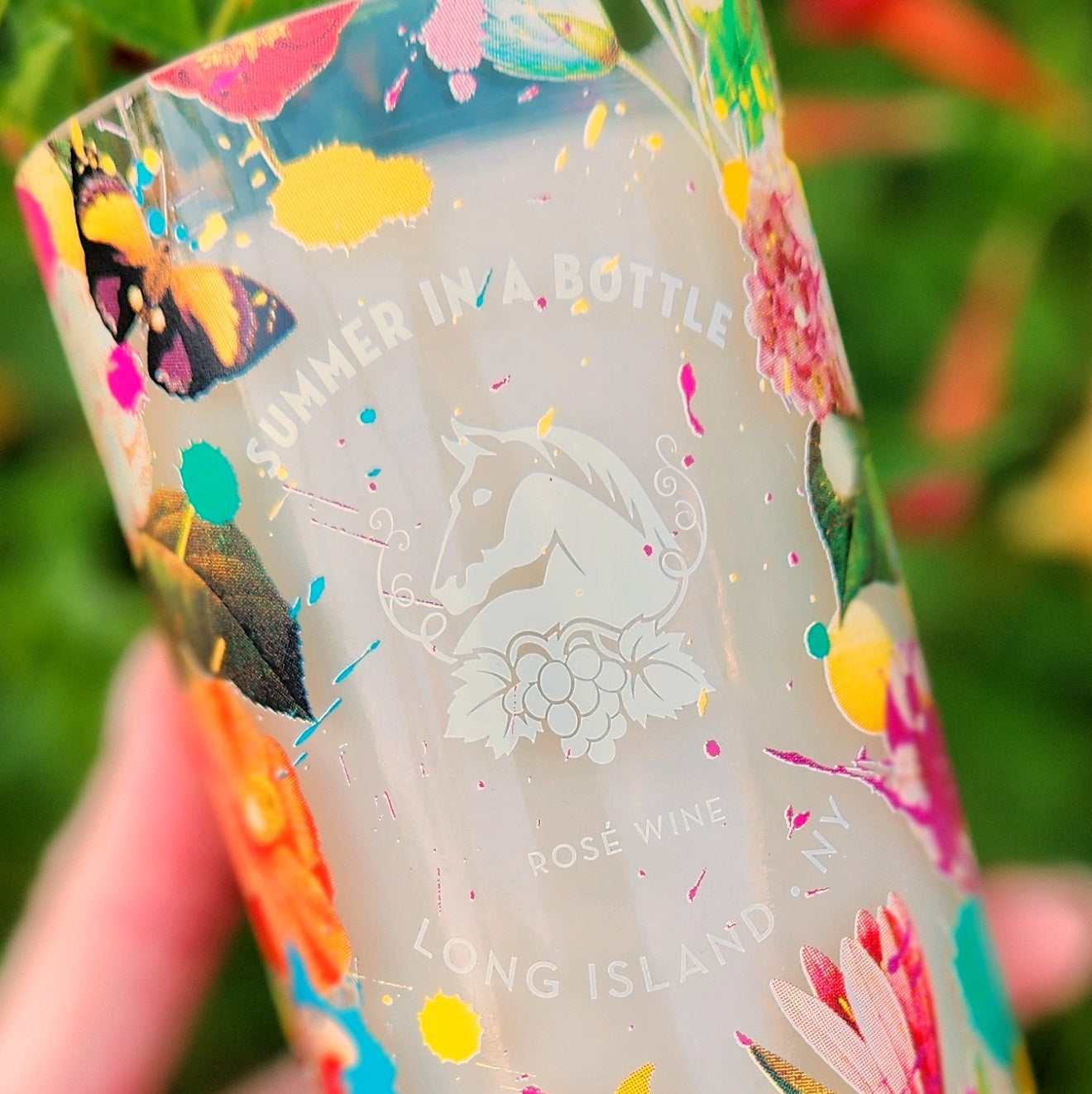Close up of “Summer in a Bottle” floral and butterfly label design, on a wine bottle candle. Held outdoors in natural light, this upcycled bottle candle glows with colorful detailing and a clean, glossy rim.