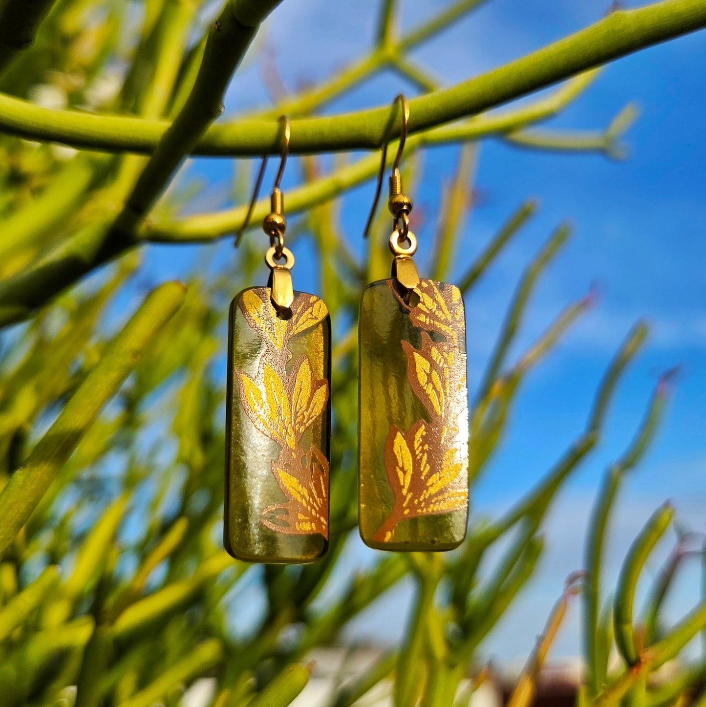 A pair of rectangular green glass drop earrings with gold-toned fishhook wires and gold leaf pattern, hanging from green branches against a blue sky background.