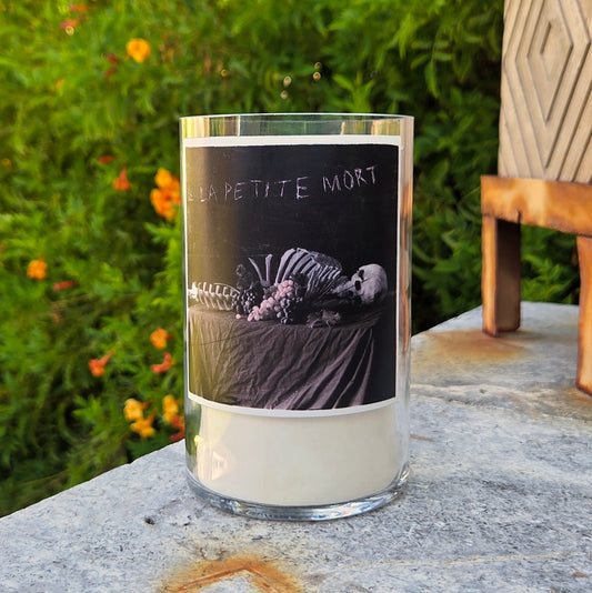 Front view of La Petite Mort wine bottle candle featuring skeleton artwork on the label, hand-poured with clean-burning coconut soy wax and wooden wick.