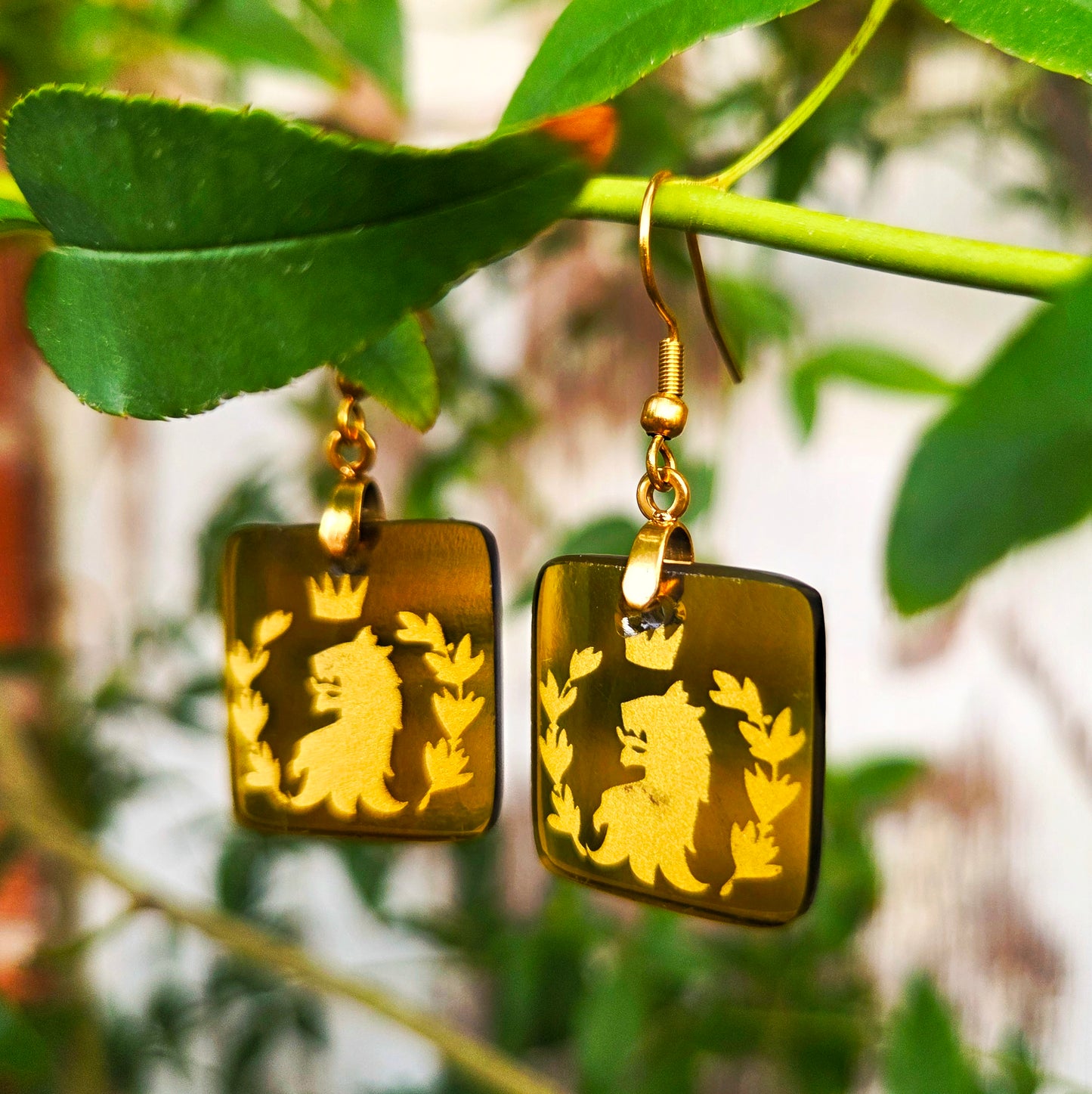 Gold-tone square drop earrings with a lion and floral motif, hanging from a green plant branch with leaves in the background.
