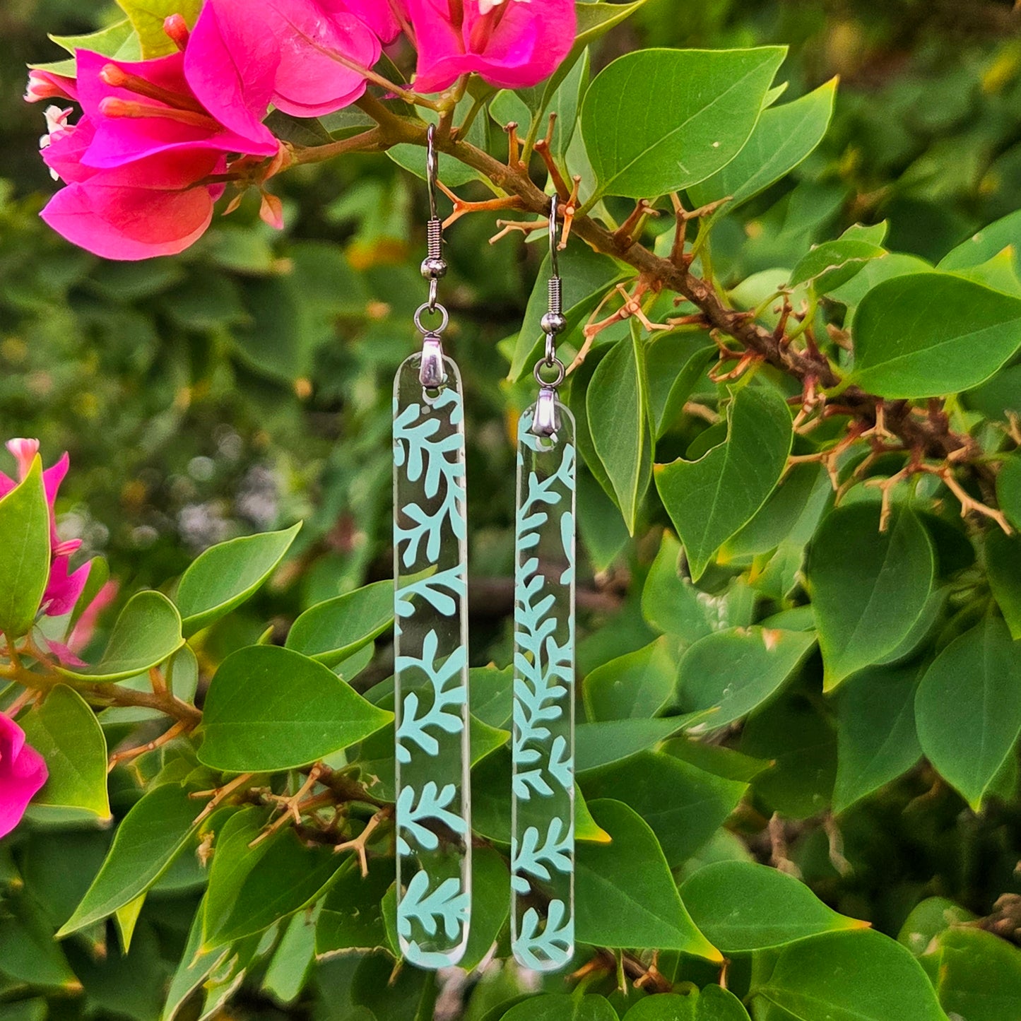 Two long rectangular clear glass drop earrings with a mint green leaf pattern, hanging from silver fishhook wires. They are displayed against a background of green leaves and bright pink bougainvillea flowers.