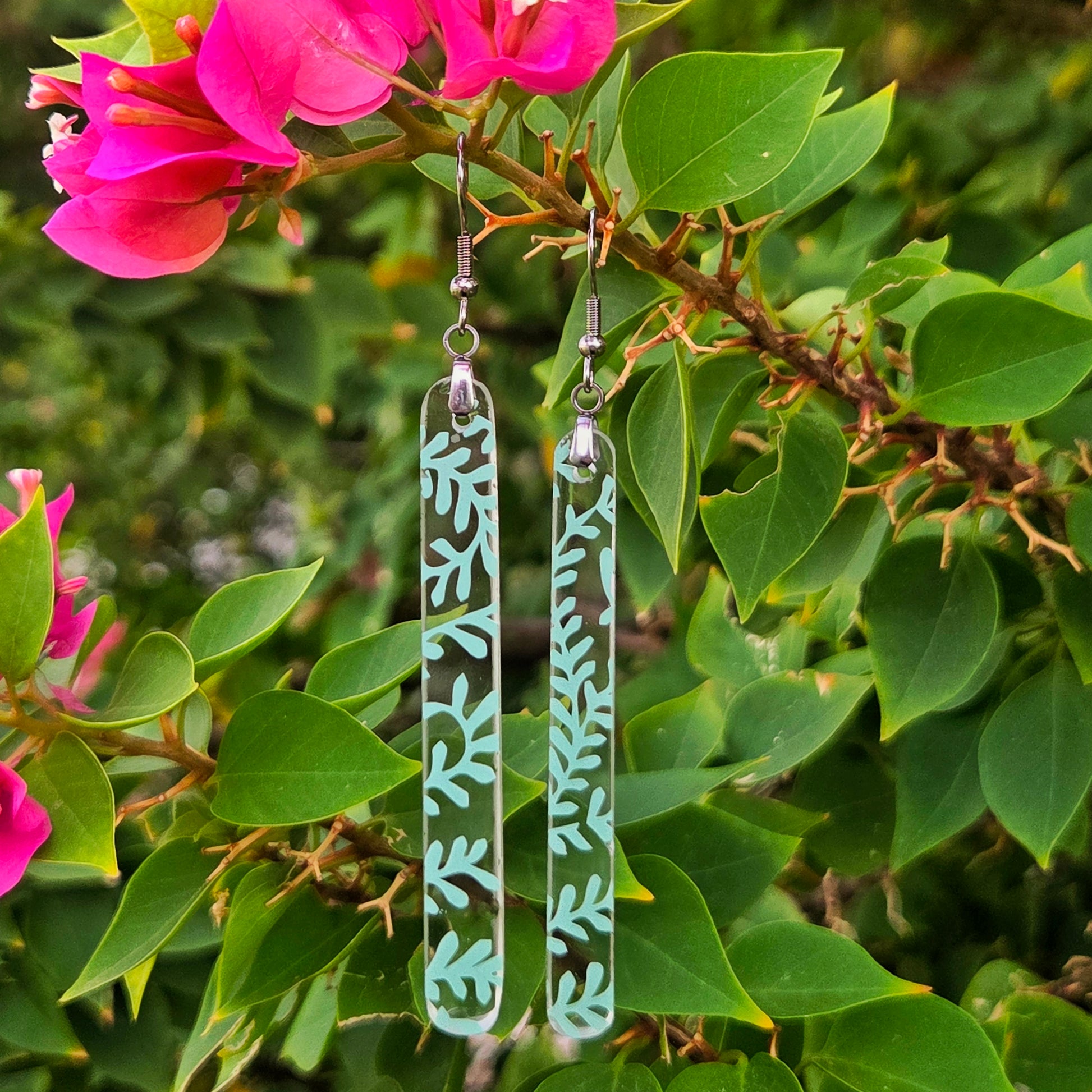 Two long rectangular clear glass drop earrings with a mint green leaf pattern, hanging from silver fishhook wires. They are displayed against a background of green leaves and bright pink bougainvillea flowers.