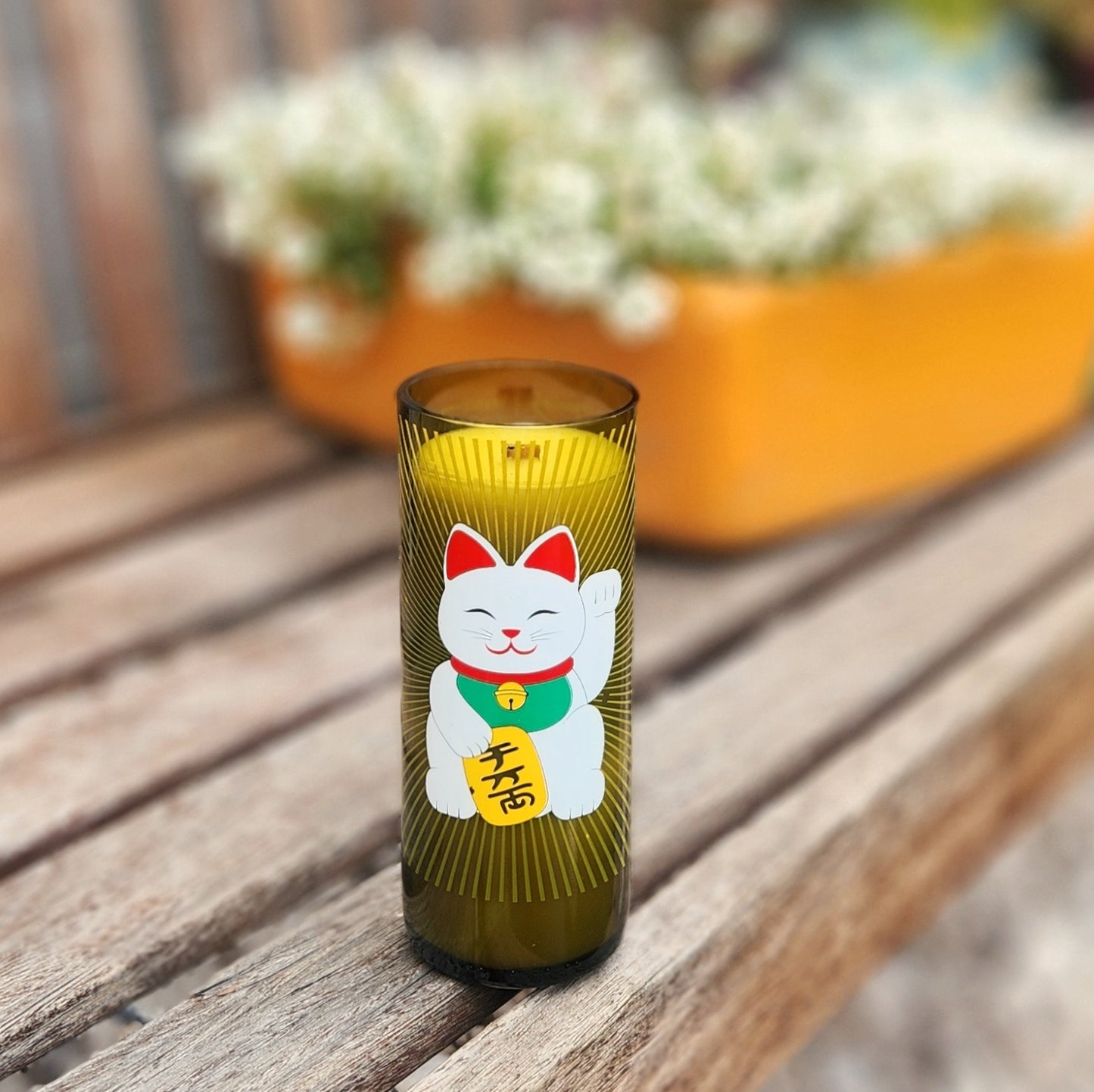 A handcrafted candle made from a recycled green wine bottle, featuring a vibrant Lucky Cat (Maneki-Neko) design. The label showcases a cheerful white cat with a raised paw, a green collar, and a gold coin with Japanese characters, set against a radiant golden sunburst pattern. The candle sits on a wooden bench outdoors, with a blurred background of blooming white flowers and a bright yellow planter, creating a warm and inviting atmosphere. The smooth glass rim and golden wax enhance its artisanal charm.