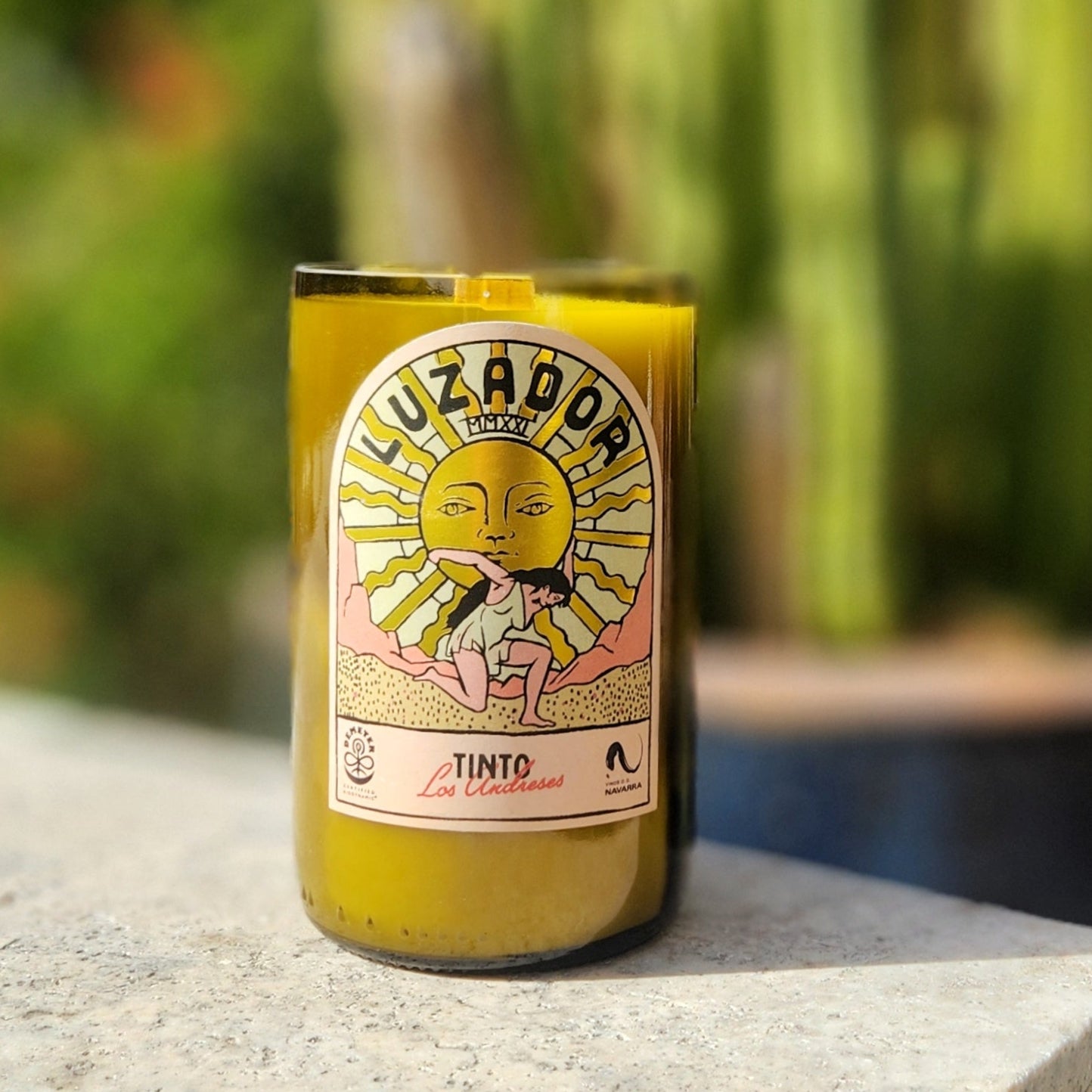 Eco-friendly candle housed in a recycled green wine bottle featuring a vivid label of a woman holding up a radiant gold sun. The hand-poured coconut soy wax and centered wooden wick add artisanal charm, while the cactus-filled desert background nods to its sustainable, Southwest-inspired roots.