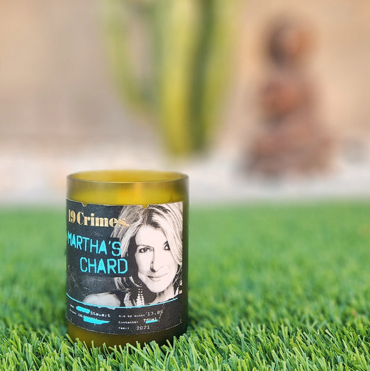 A hand-poured candle crafted from coconut-soy wax, housed in a recycled green wine bottle with a smooth, frosted rim. The label features a design from "19 Crimes Martha's Chard," showcasing a portrait of Martha Stewart in bold, modern typography. The candle is placed on a lush patch of artificial grass, with a blurred background of a cactus and a decorative sculpture, adding a stylish and eco-friendly vibe to the scene. Perfect for upcycled wine bottle decor.