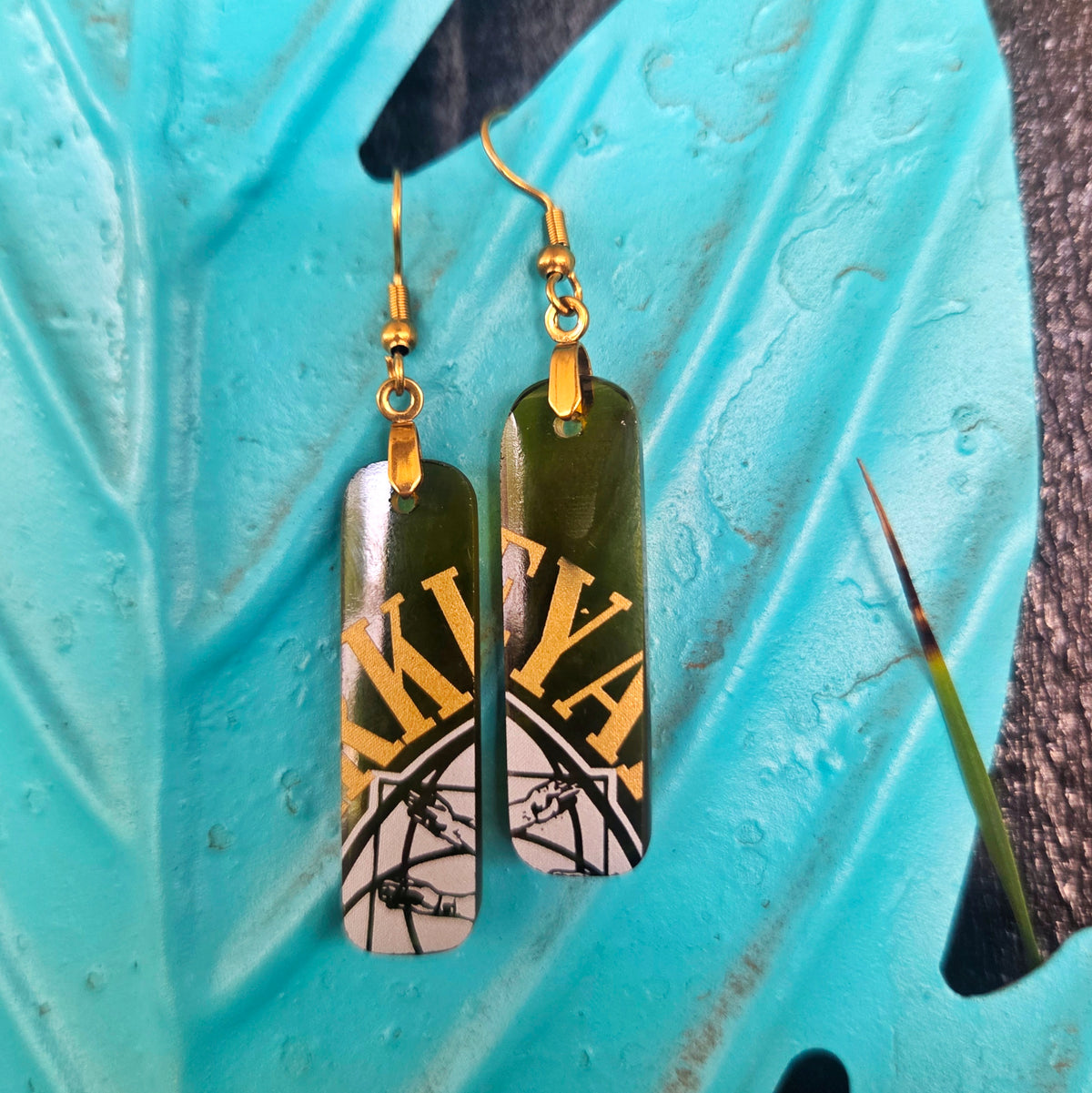 A pair of rectangular drop earrings with gold fishhook wires, featuring a dark olive green top section with gold lettering and a white bottom section with black graphic details, displayed on a textured turquoise leaf.