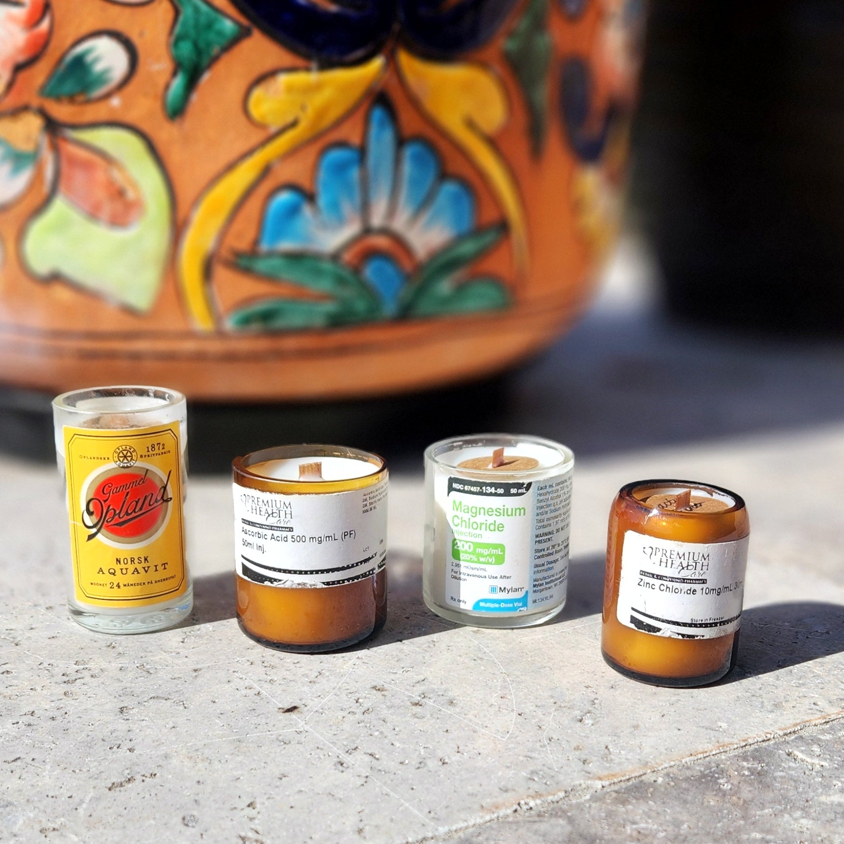 A collection of four upcycled candles, each crafted in small recycled glass containers, including repurposed bottles and pharmaceutical jars. The containers feature original or vintage labels, such as "Gammel Opland Norsk Aquavit" and "Magnesium Chloride," adding unique character and charm. Each candle is hand-poured with coconut-soy wax and includes a wooden wick. The set is displayed outdoors on a concrete surface, with a vibrant, floral-patterned ceramic pot in the background, creating a bright and color