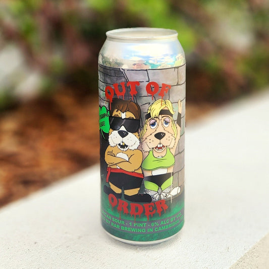 A recycled beer can candle featuring a cartoon label inspired by Mortal Kombat. The design shows two anthropomorphic dog characters dressed like 90s fighters—one muscular and wearing sunglasses, the other in a crop top and ponytail—standing in front of a brick wall with bold red lettering that says “OUT OF ORDER.” The can sits on a smooth outdoor ledge with blurred greenery in the background. 