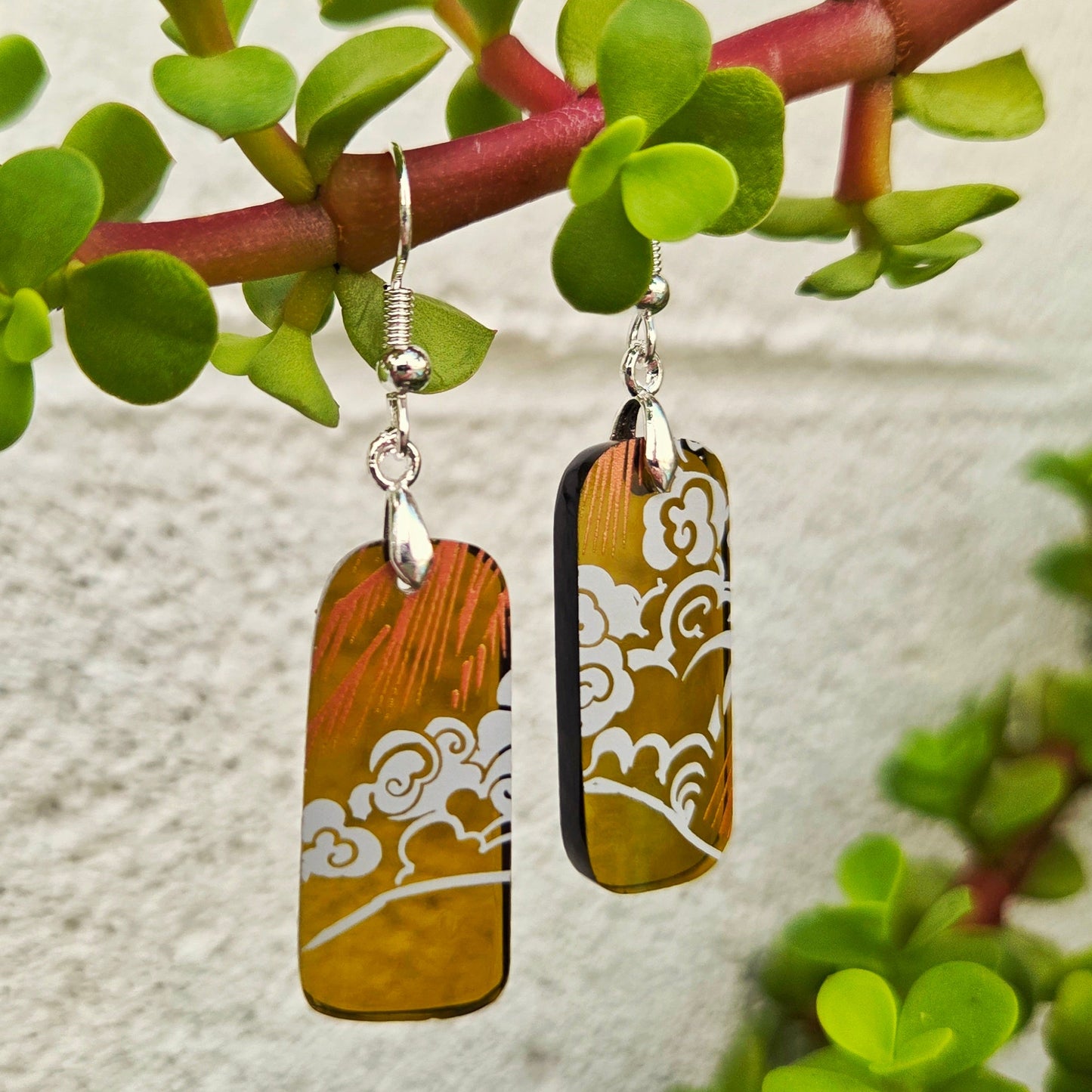 A pair of rectangular drop earrings with a marbled amber, orange, and brown design featuring white cloud-like patterns, hanging from silver fishhook wires. The earrings are displayed hanging from a green succulent branch against a textured white wall.