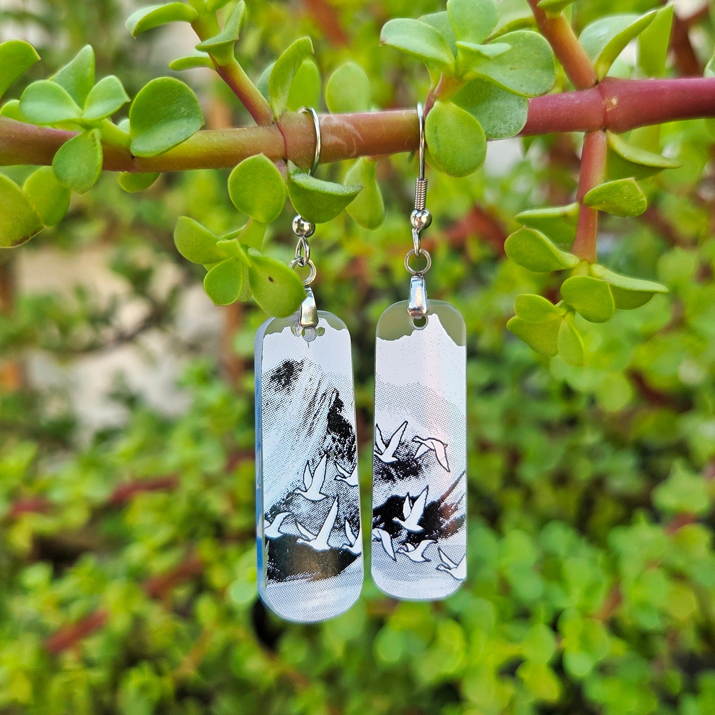 A pair of rectangular glass drop earrings with a black and white abstract landscape print, hanging from silver fishhook wires on a green plant branch with foliage in the background.