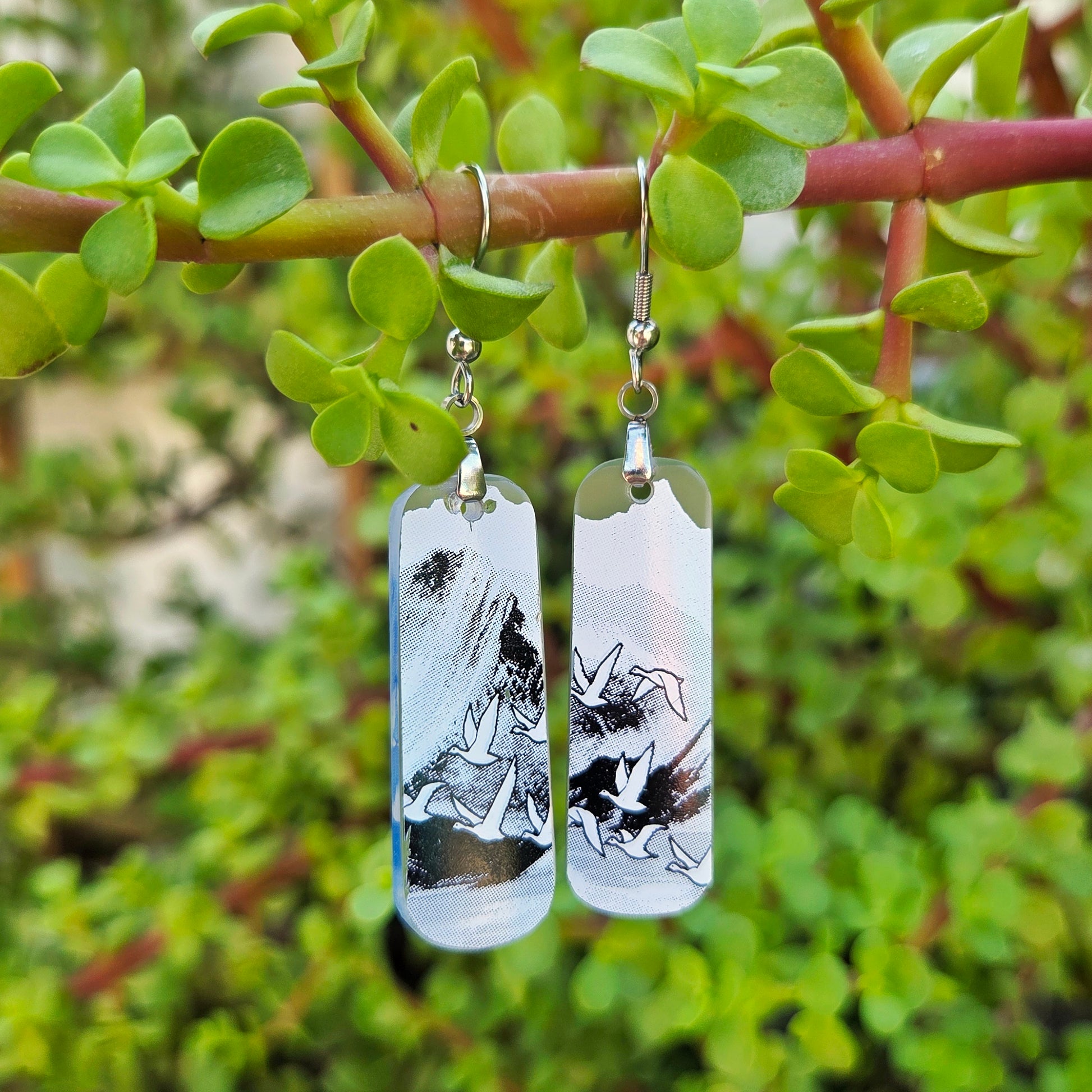 A pair of rectangular glass drop earrings with a black and white abstract landscape print, hanging from silver fishhook wires on a green plant branch with foliage in the background.