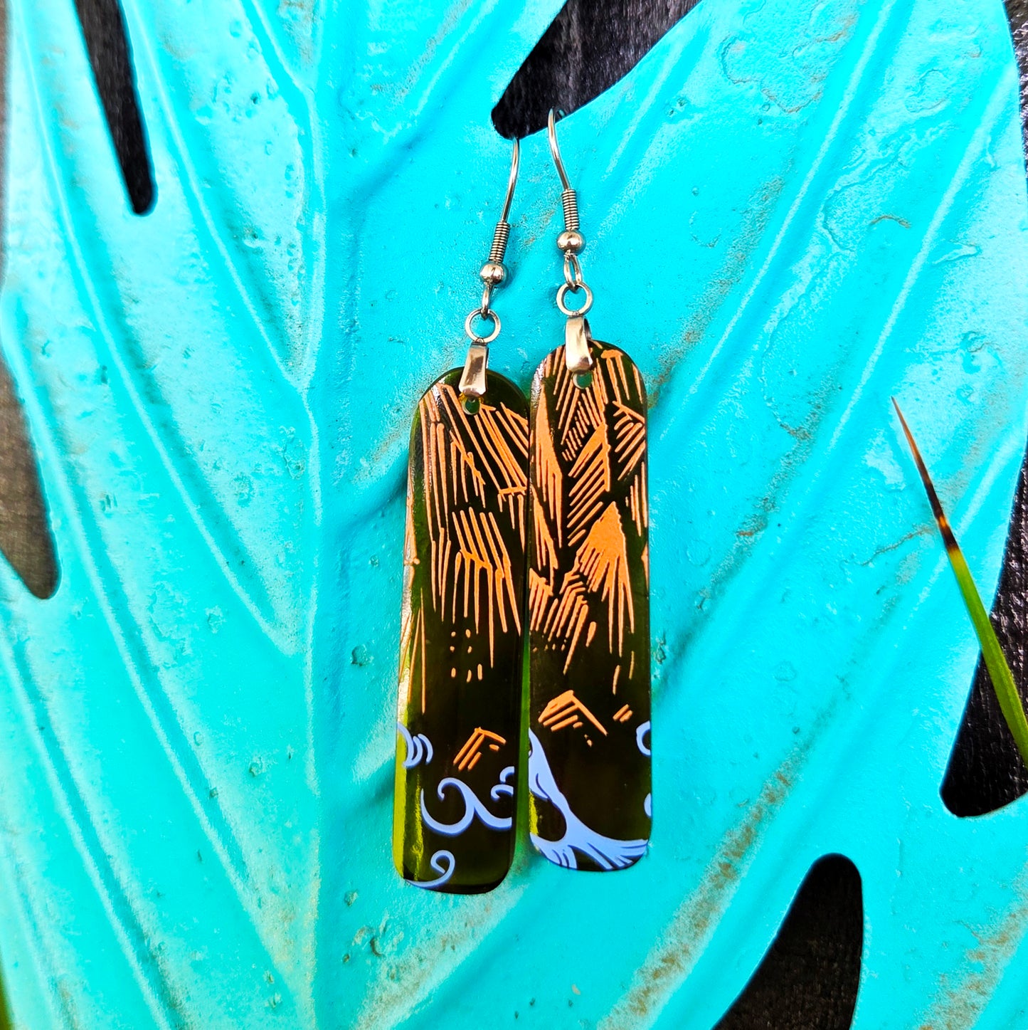 A pair of rectangular drop earrings with a printed ocean and mountain scene, hanging against a turquoise leaf-shaped background.