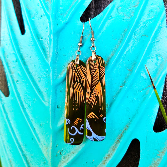 A pair of rectangular drop earrings with a printed ocean and mountain scene, hanging against a turquoise leaf-shaped background.