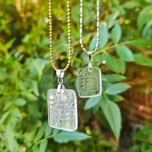 Two clear glass necklaces with music clef patterning hanging against a green leafy background