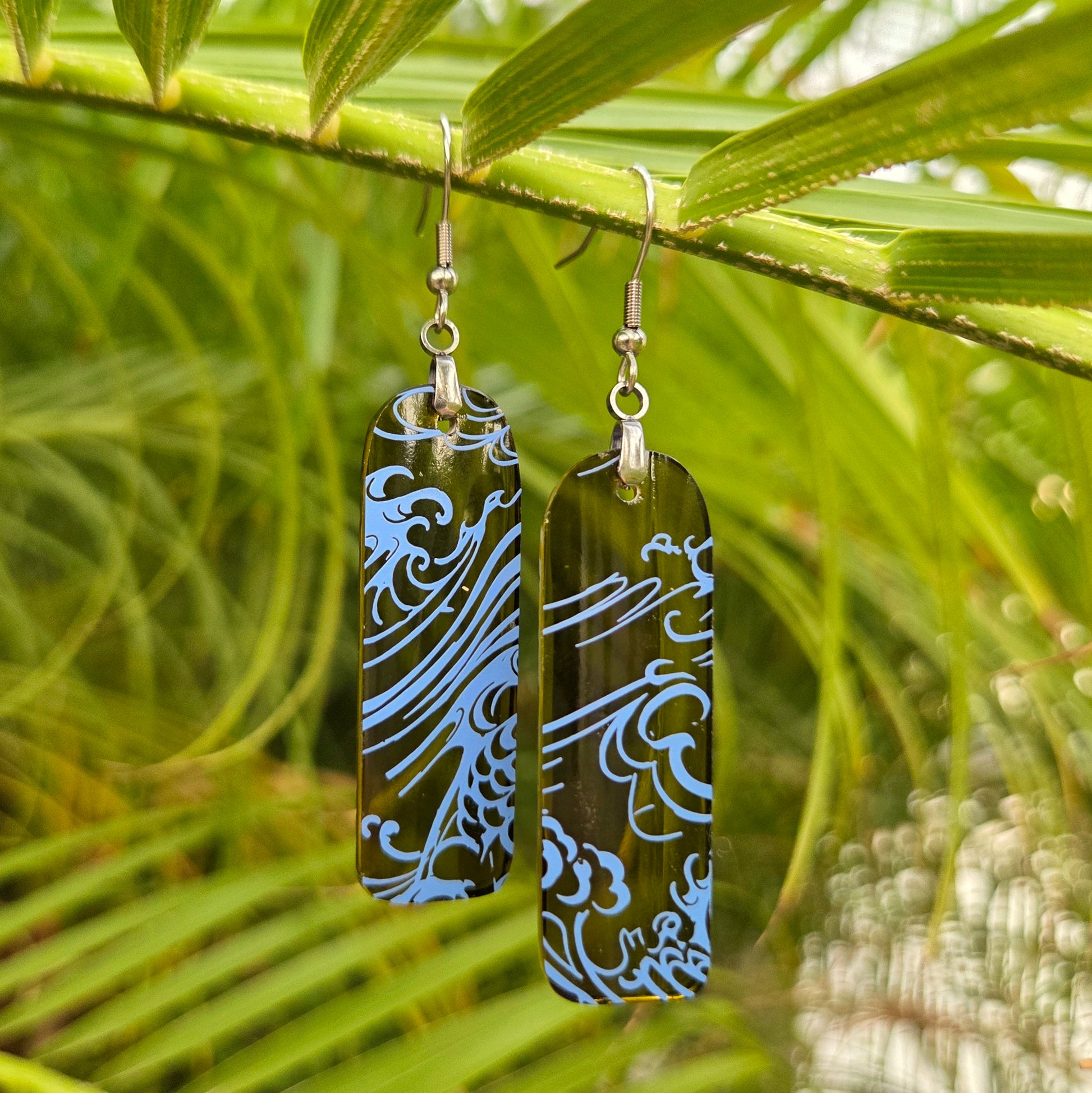 Two rectangular drop earrings with a dark green translucent background and light blue wave patterns, hanging from silver fishhook wires against a blurred background of green palm leaves.