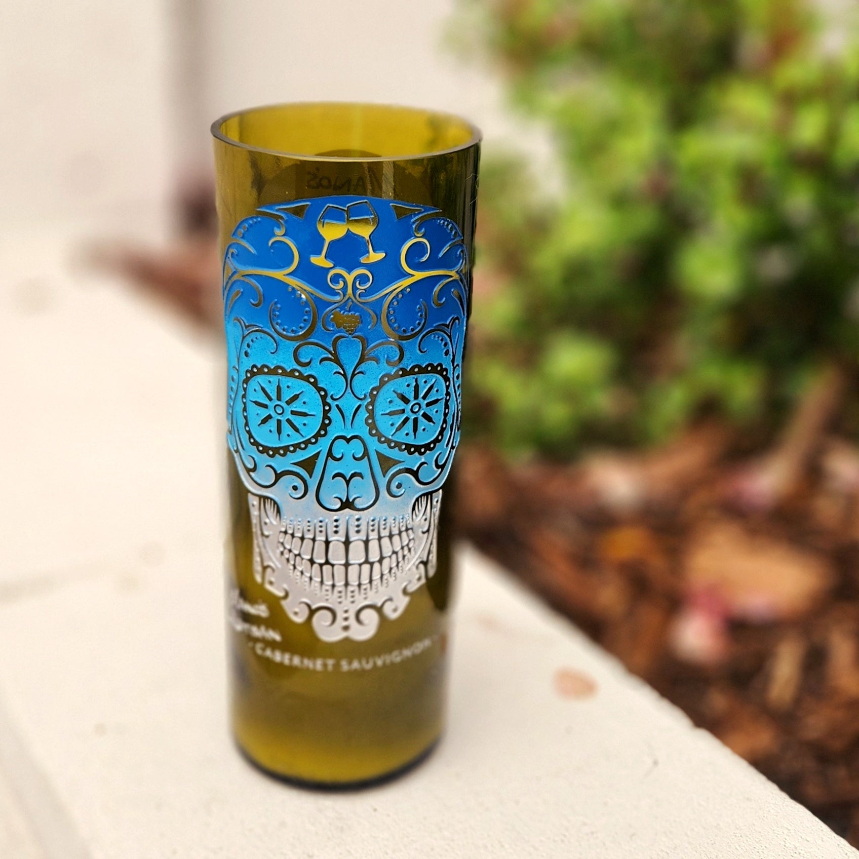 A hand-poured eco-friendly wine bottle candle made from a recycled bottle, featuring a vibrant sugar skull design in ombre blue and silver. The coconut soy wax and clean-burning wooden wick are housed in smooth, polished glass, giving this artistic upcycled candle a bold, Día de los Muertos-inspired aesthetic.

