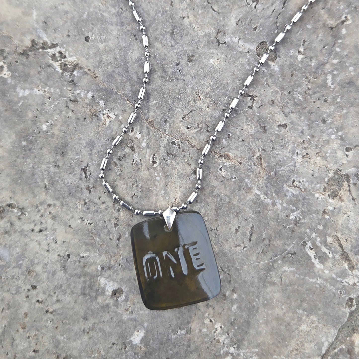 Necklace with a glass dog tag engraved with 'ONE' on a stone surface