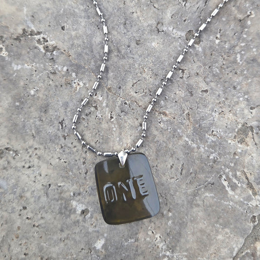 Necklace with a glass dog tag engraved with 'ONE' on a stone surface