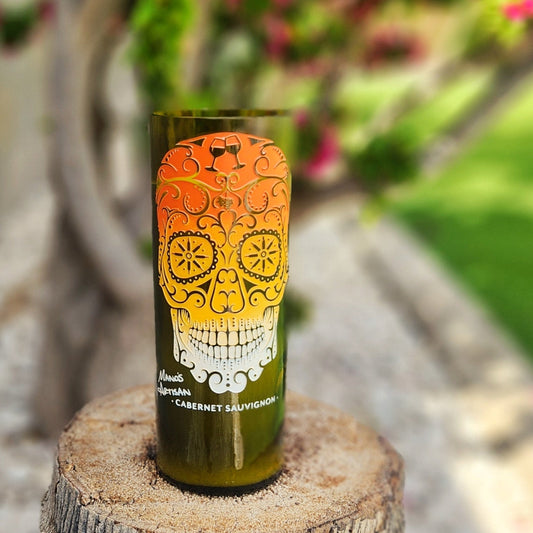  A hand-poured coconut soy candle in a recycled wine bottle, featuring an intricate sugar skull design with an orange-to-white gradient. The eco-friendly wine bottle candle has a polished rim and a wooden wick, creating a sustainable yet artistic touch. Placed on a rustic tree stump with lush greenery and vibrant pink flowers in the background, this Día de los Muertos-inspired candle blends bold aesthetics with sustainability.