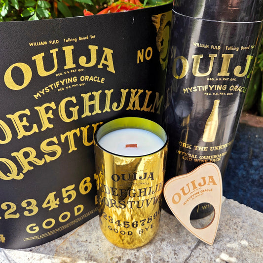 Gold wine bottle featuring a ouija board made into a candle setting in front of an actual ouija board.