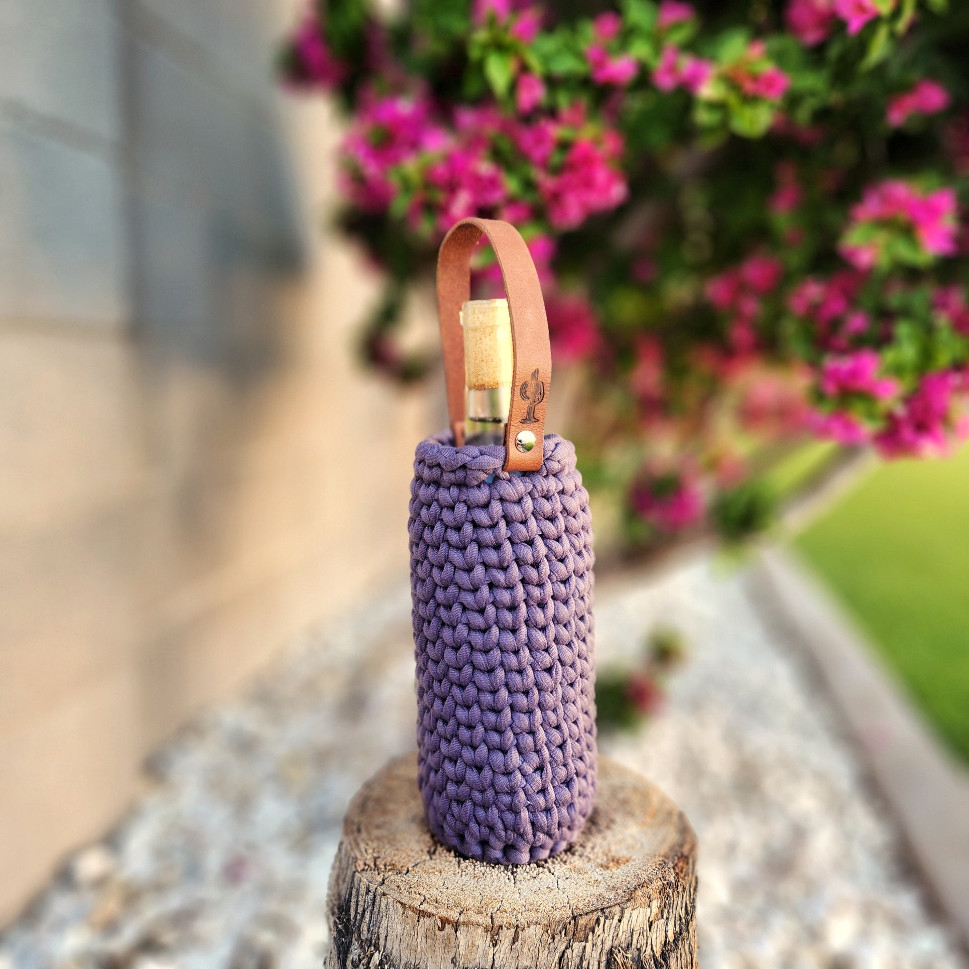 A hand-poured eco-friendly wine bottle candle snugly wrapped in a thick, pewter purple crocheted carrier with a leather handle featuring a stamped cactus design. The candle rests on a tree stump with vibrant pink flowers and lush greenery in the blurred background.