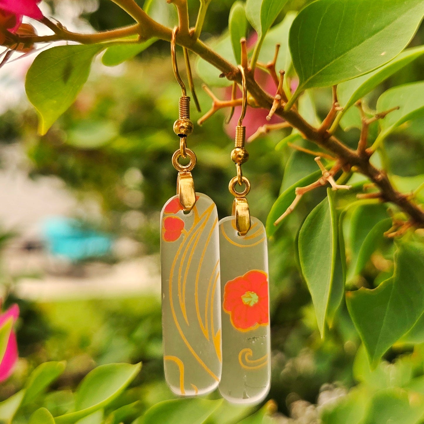 Two rectangular glass dangle earrings with gold floral and wave patterns, hanging from gold hooks against a background of green leaves and pink flowers.