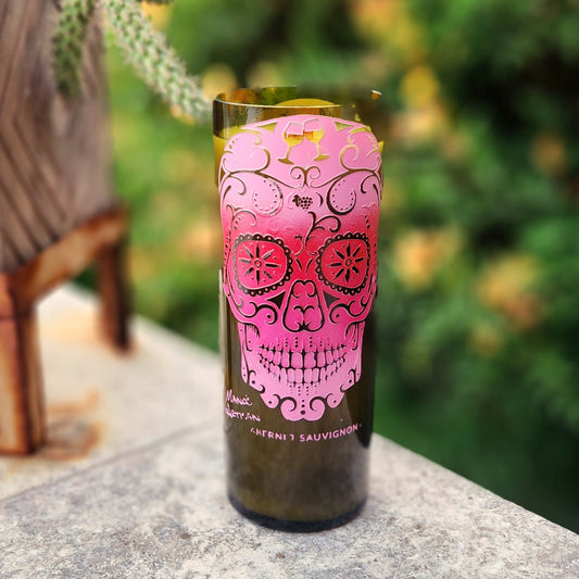 An eye-catching eco-friendly wine bottle candle featuring a vivid pink sugar skull design etched with decorative details and wine glasses. The candle is hand poured into a recycled bottle, filled with coconut soy wax and topped with a wooden wick for a clean, crackling burn.