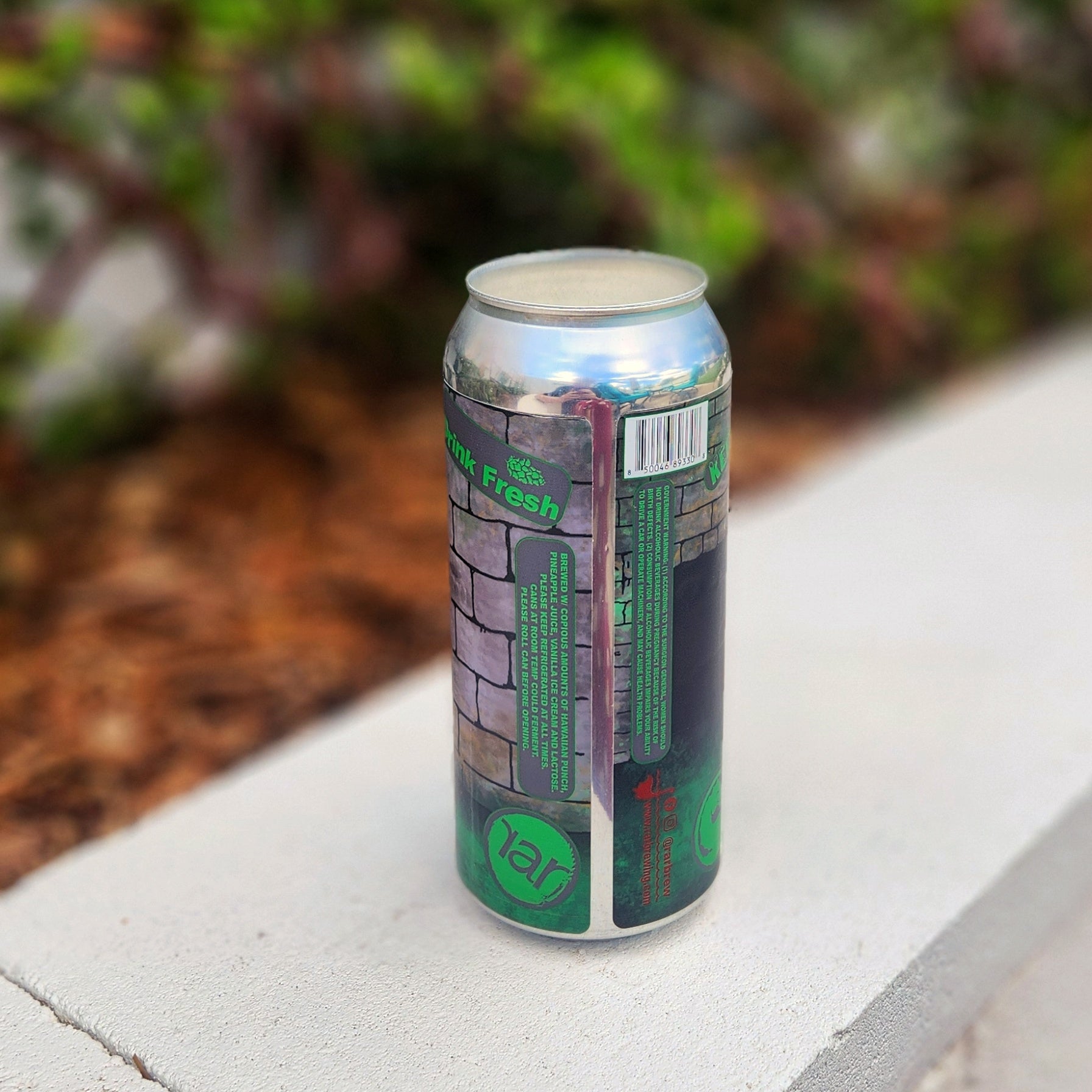 Back view of a recycled beer can candle featuring a Mortal K-inspired design by RaR Brewing. The label wraps around the silver can with a cartoon-style stone wall background and bold green and red text, including phrases like “Drink Fresh” and brewery details. The candle inside has a creamy white wax and wooden wick. The can rests on a pale concrete ledge with mulch and blurred greenery in the background.