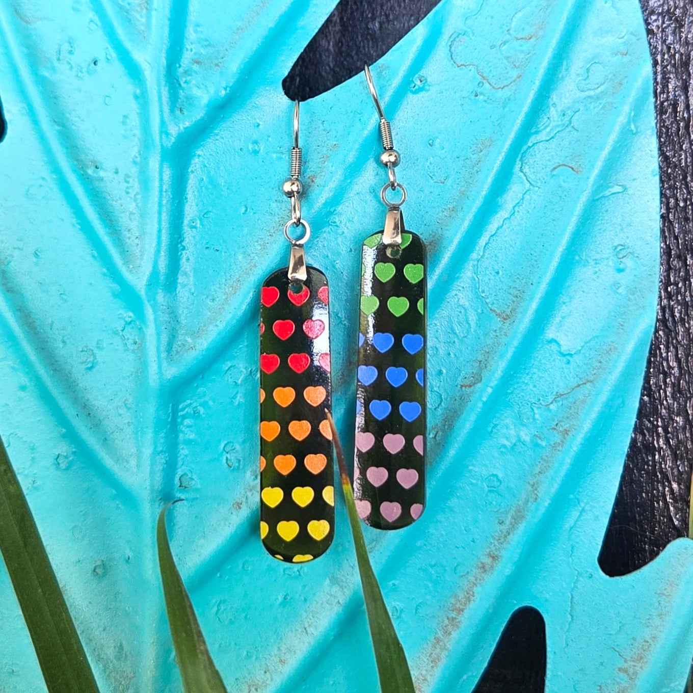 Colorful glass wine bottle earrings with a rainbow heart design on a decorative leaf with a dark background