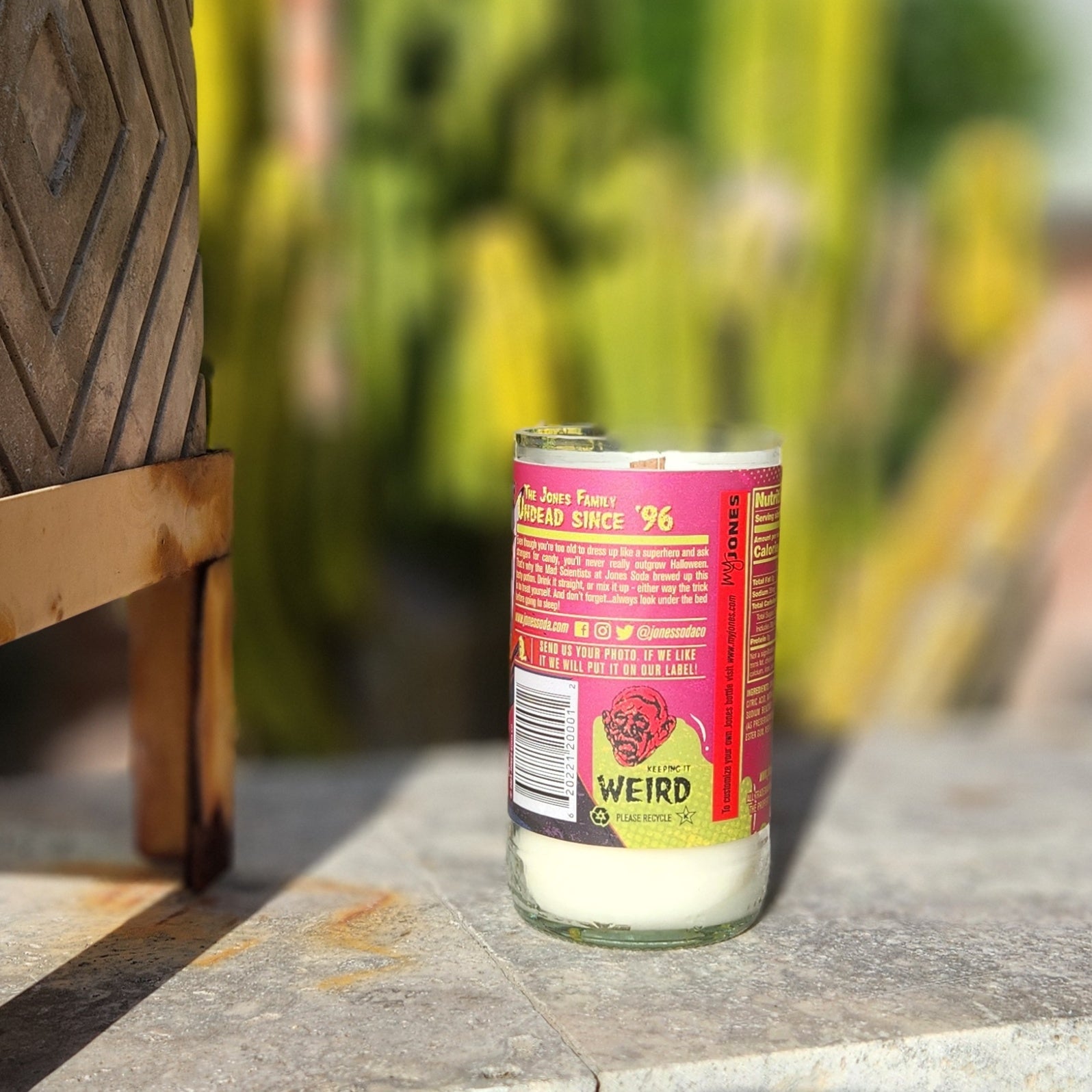 A recycled soda bottle candle featuring a bright red and yellow vampire-themed label that reads “Undead Since ‘96” with quirky branding details from Jones Soda; the eco-friendly wine bottle candle sits on stone in bright natural light with cactus blurred in the background.