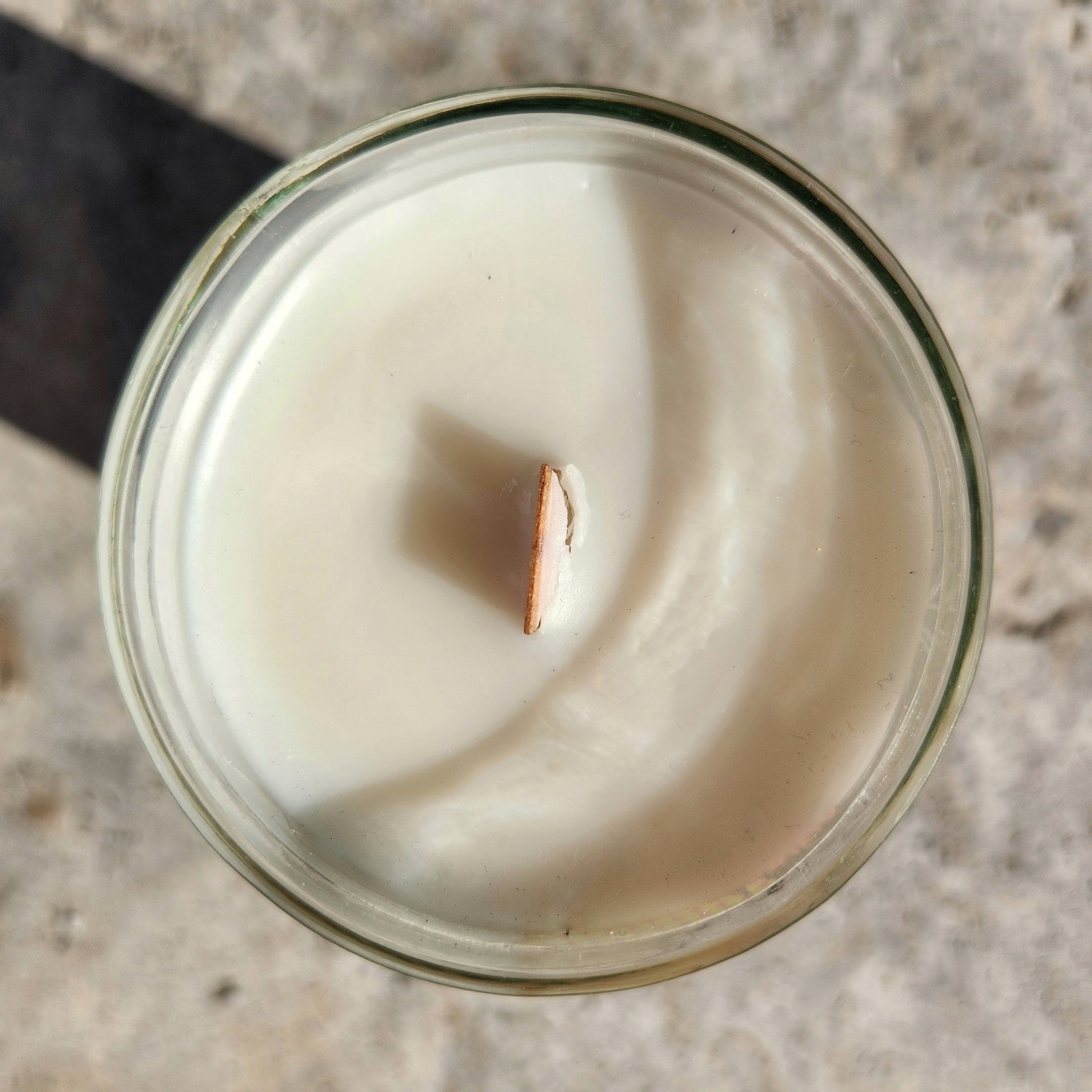 A direct top view of an unlit coconut soy wine bottle candle with a wooden wick centered in smooth white wax, housed in a recycled bottle with clean edges and a faint green tint to the glass.