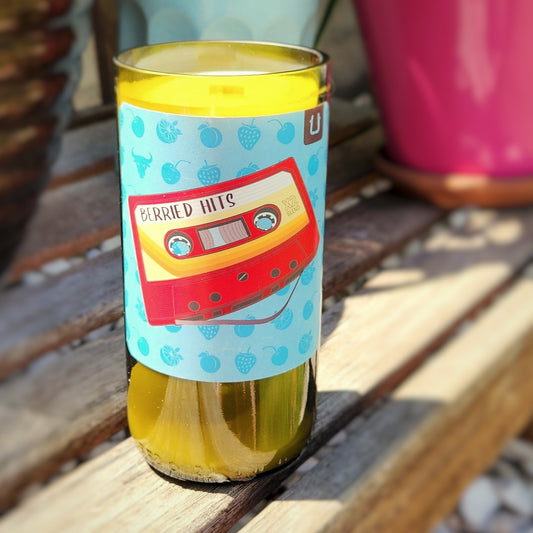 Hand-poured recycled wine bottle candle with a vibrant label featuring a retro cassette tape design labeled 'Berried Hits,' set against a colorful background with berry illustrations, placed on a wooden surface with potted plants in the background.