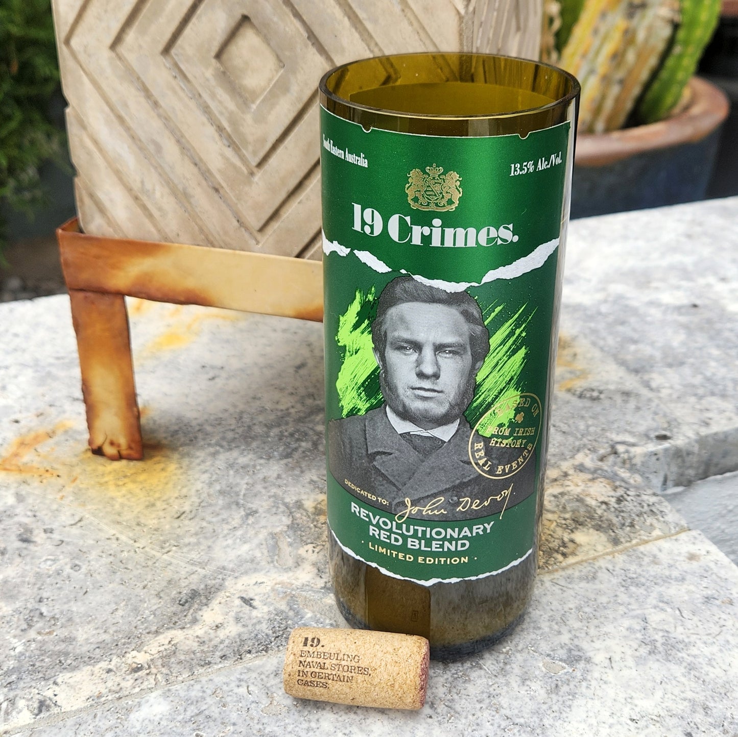  A hand-poured coconut soy candle in a recycled 19 Crimes Revolutionary Red Blend wine bottle, featuring John Devoy on the green label. The eco-friendly wine bottle candle has a smooth, polished rim and a wooden wick. A branded cork with "19" and historical text rests in front. Set on a stone surface with a modern planter and cacti in the background, this upcycled wine bottle candle blends sustainability with bold design.
