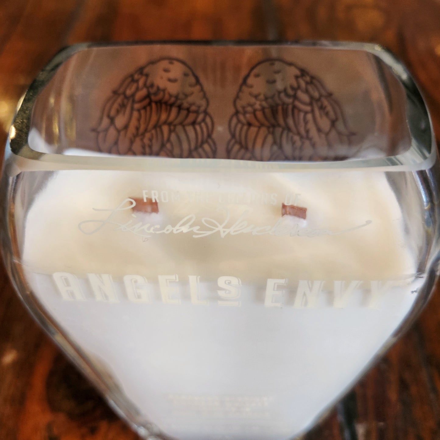 Close-up of a double wooden wick eco-friendly candle housed in a recycled Angel’s Envy liquor bottle, highlighting the creamy coconut soy wax and wing design visible through the glass.


