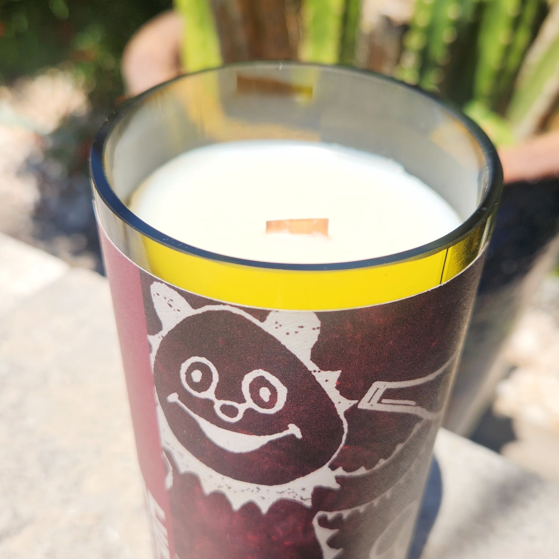 Close-up side view of a clean-burning recycled wine bottle candle, highlighting the glossy rim and white coconut soy wax with a wooden wick; playful grape design visible on the label.

