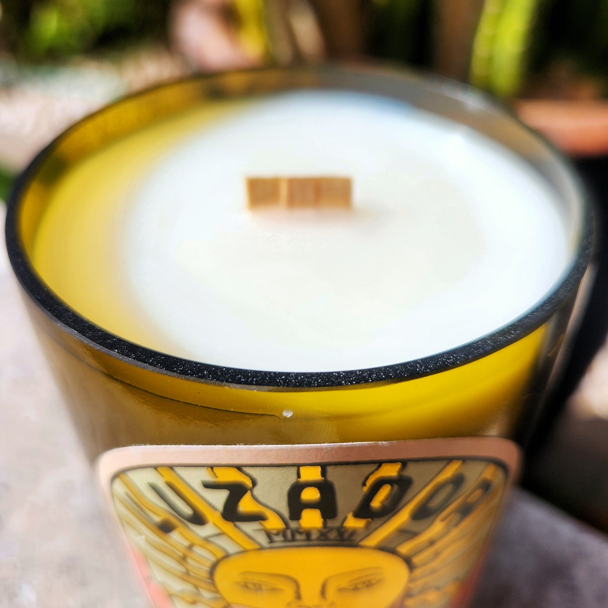 A close-up of the hand-poured coconut soy candle in a recycled Luzador wine bottle, showcasing the smooth, glossy rim and centered wooden wick. The clean edge highlights its upcycled craftsmanship, while the sunny label peeks through in the background, adding a vibrant, artisanal touch.
