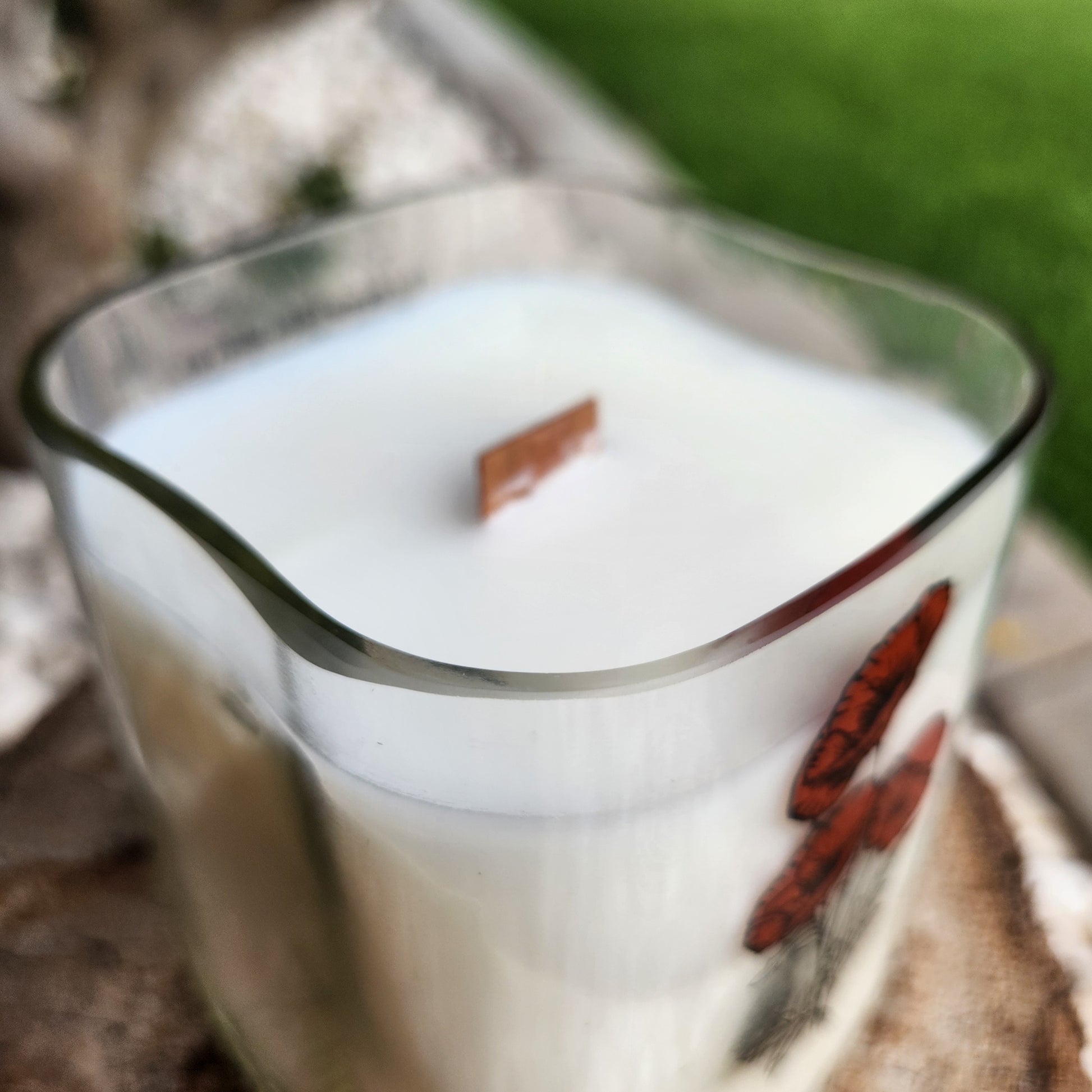 Alt text: Close-up side view of a coconut soy wax candle with a wooden wick, housed in a curved, recycled milk bottle. The clear glass rim showcases a smooth finish, emphasizing the candle’s handmade, non-toxic design.

